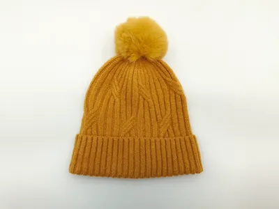 Leuchtend senfgelbe Wintermütze aus Zopfstrick mit großem Kunstfell-Bommel und Umschlagbündchen auf weißem Hintergrund