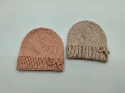 Zwei weiche Rippstrickmützen mit Schleifenverzierung in Rosa und Beige, nebeneinander auf hellem Untergrund