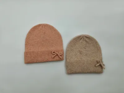 Zwei feine Strickmützen mit Rippbündchen und kleiner Schleifenverzierung in Blush-Rosa und Beige auf weißem Hintergrund