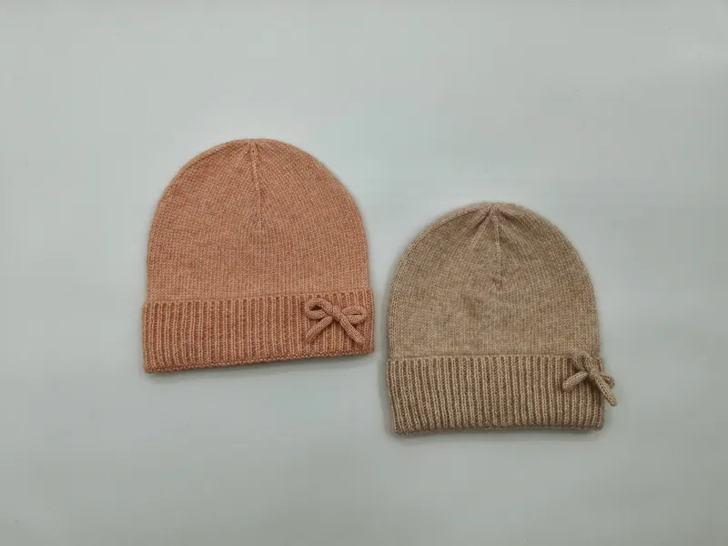 Zwei feine Strickmützen mit Rippbündchen und kleiner Schleifenverzierung in Blush-Rosa und Beige auf weißem Hintergrund