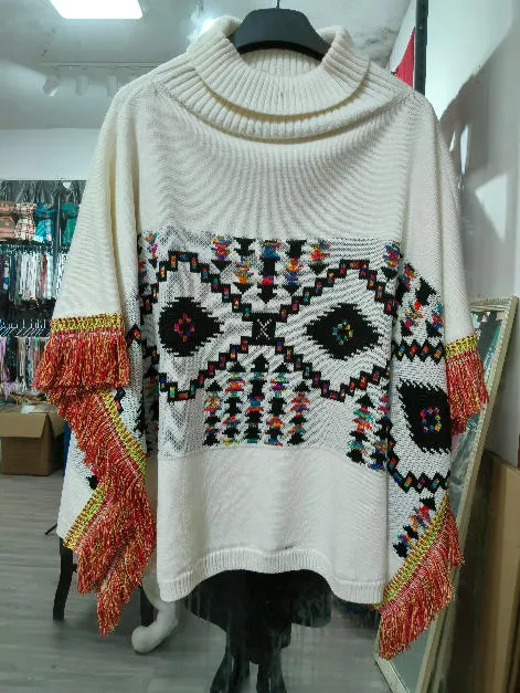 Cremefarbener Oversize-Rollkragenponcho mit buntem geometrischem Aztekenmuster und roten Fransen an den Ärmeln auf Schneiderpuppe