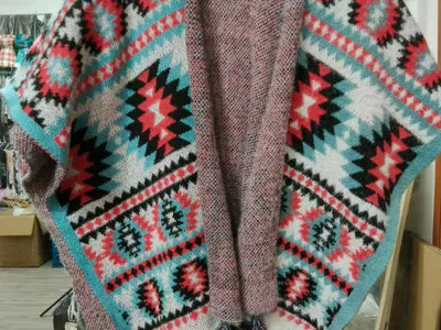 Offener Strickponcho mit Aztekenmuster in Rot, Petrol, Schwarz und Weiß sowie bunten Quastenfransenabschlüssen auf Kleiderbügel