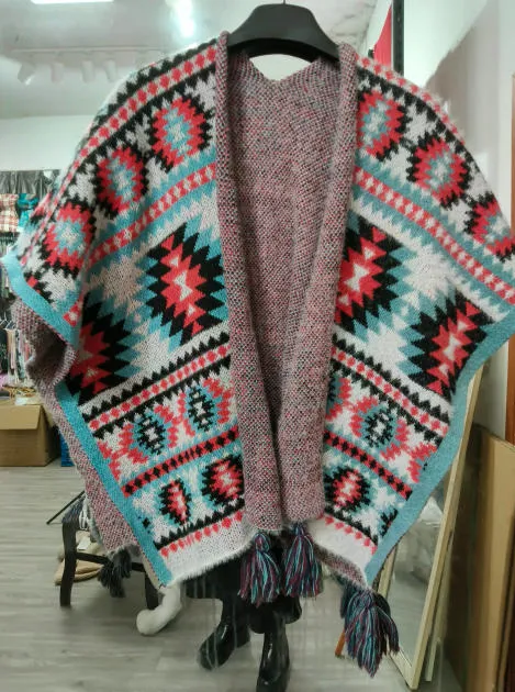 Offener Strickponcho mit Aztekenmuster in Rot, Petrol, Schwarz und Weiß sowie bunten Quastenfransenabschlüssen auf Kleiderbügel