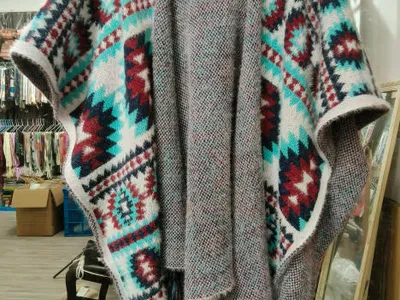 Offener Strickponcho mit geometrischem Aztekenmuster in Petrol, Burgund und Weiß mit Quastenfransensaum auf Kleiderbügel
