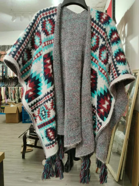 Offener Strickponcho mit geometrischem Aztekenmuster in Petrol, Burgund und Weiß mit Quastenfransensaum auf Kleiderbügel