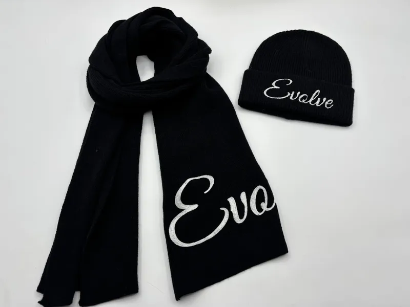 Schwarzes Rippenstrick-Winterset aus Schal und Beanie-Mütze mit weißem bestickten Kursivschrift-Aufdruck auf weißem Hintergrund