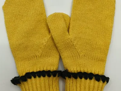 Paar senfgelbe Strickfäustlinge mit Rippenbündchen und schwarzen dekorativen Bommel-Details an den Manschetten