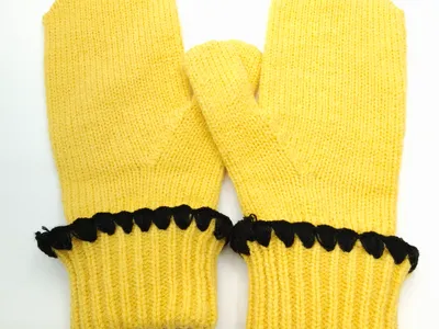 Paar gelbe Strickfäustlinge mit schwarz gehäkelter Bogenkante entlang des gerippten Bündchens, flach auf weißem Hintergrund