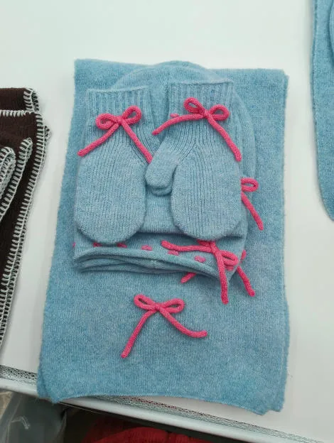 Hellblaues gestricktes Babywinter-Geschenkset mit Schal, Mütze und Fäustlingen mit rosafarbenen Schleifen-Applikationen, flach drapiert