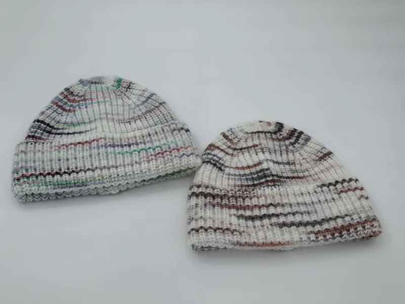Zwei handgestrickte Rippstrick-Beanies in Weiß mit farbigen Streifenmustern – eine in kühlen Tönen, eine in warmen Erdfarben, auf weißem Hintergrund