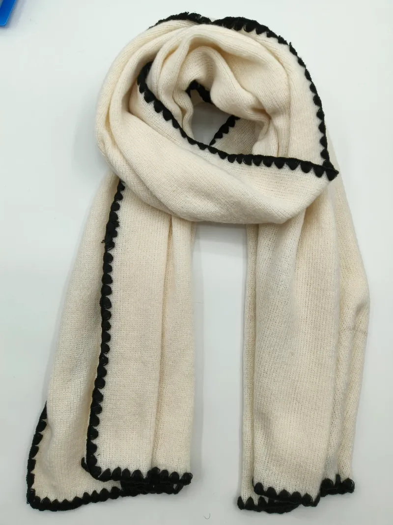 Elfenbeinfarbener gestrickter Winterschal mit dekorativer schwarzer Bogenkante, flach auf hellem Untergrund drapiert