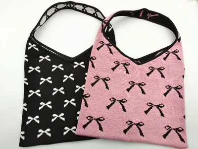 Zwei gestrickte Hobo-Bags mit Allover-Schleifenmuster: eine schwarz mit weißen Schleifen, eine pink mit schwarzen Schleifen, mit Schulterriemen