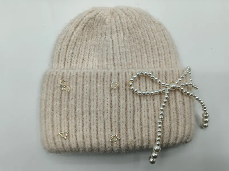 Gestrickte Wintermütze in Creme mit feiner Rippenstruktur, verziert mit einer Perlenschleife und funkelnden Strass-Akzenten am Umschlag
