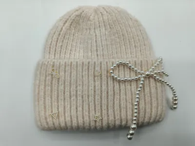 Gestrickte Wintermütze in Creme mit feiner Rippenstruktur, verziert mit einer Perlenschleife und funkelnden Strass-Akzenten am Umschlag