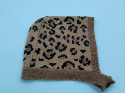 Gestrickte Säuglingsbonnetmütze im Leopardenprint mit beigem Hintergrund und schwarzen Flecken, mit Rippenrand und Kinnbändchen – flach auf weißem Untergrund