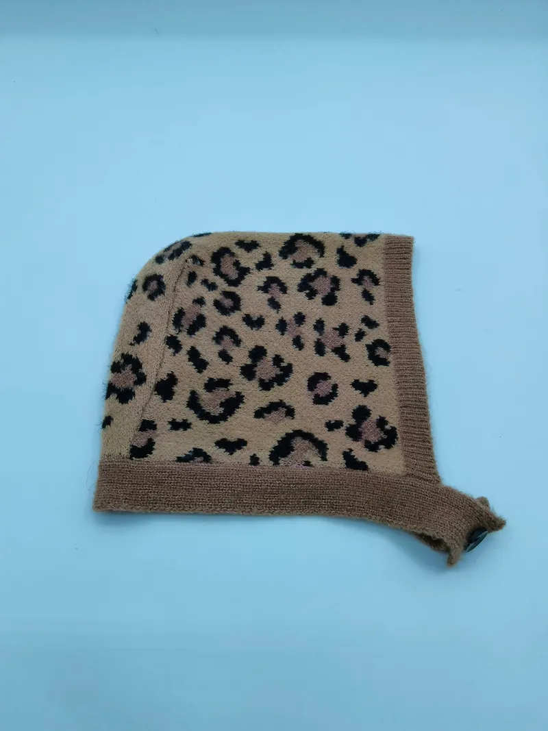Gestrickte Säuglingsbonnetmütze im Leopardenprint mit beigem Hintergrund und schwarzen Flecken, mit Rippenrand und Kinnbändchen – flach auf weißem Untergrund