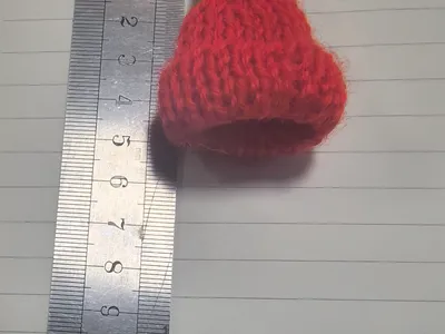 Winzige handgestrickte Wollmütze in Rot mit grünem Bommelpunkt, neben einem Lineal mit ca. 4 cm Höhe