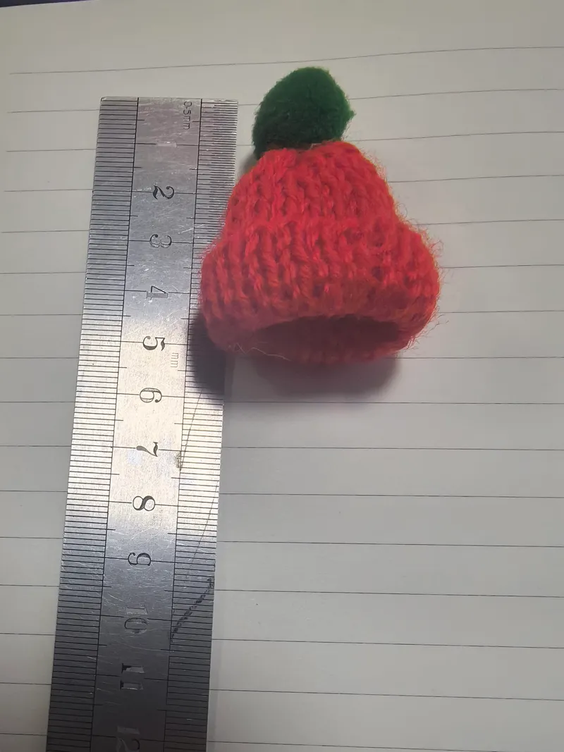 Winzige handgestrickte Wollmütze in Rot mit grünem Bommelpunkt, neben einem Lineal mit ca. 4 cm Höhe