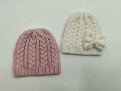 Zwei Wintermützen mit klassischem Zopfstrickmuster – eine in gedämpftem Rosa, eine in Cremeweiß mit seitlicher Schleifenverzierung