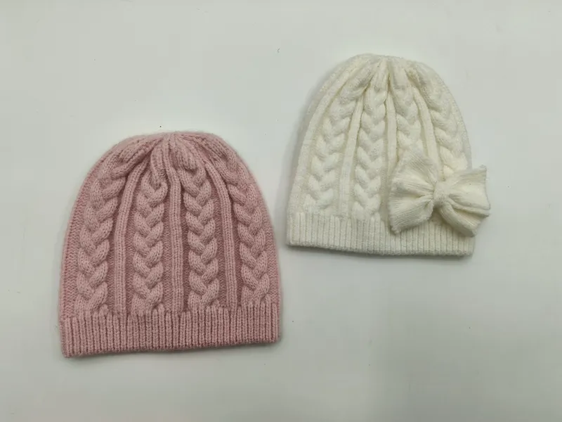 Zwei Wintermützen mit klassischem Zopfstrickmuster – eine in gedämpftem Rosa, eine in Cremeweiß mit seitlicher Schleifenverzierung