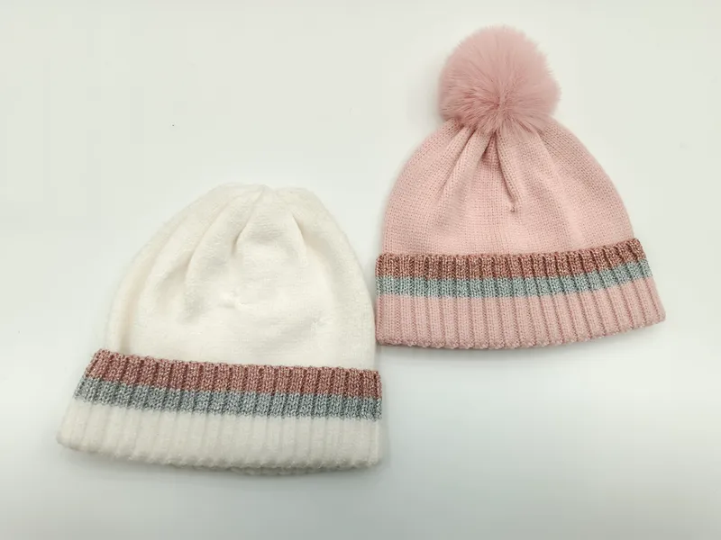 Zwei Rippenstrick-Beanies in Weiß und Rosa mit schimmernden Metallic-Streifen am Umschlag – pinke Mütze mit Kunstfell-Bommel obenauf