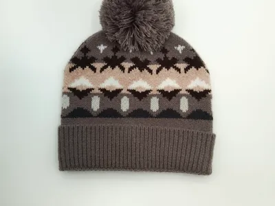 Grau gestrickte Wintermütze mit Fair-Isle-Motiven in Rosa, Braun und Weiß sowie flauschigem Bommel auf der Spitze, Studioaufnahme