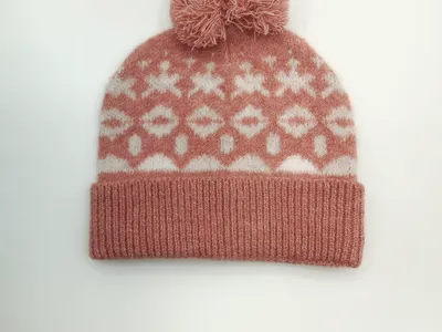 Rosafarbene Strickmütze mit weißen geometrischen Schneeflocken-Ornamenten und flauschigem Bommel, Produktfoto auf hellem Hintergrund