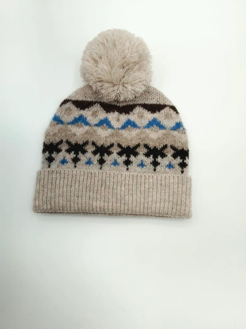 Haferfarben gestrickte Mütze mit blau-schwarzem Schneeflocken-Jacquardmuster und großem cremefarbenem Bommel auf der Kuppe