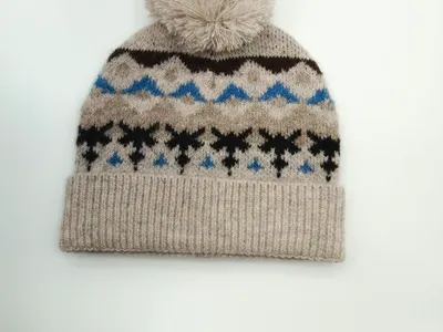 Haferfarben gestrickte Mütze mit blau-schwarzem Schneeflocken-Jacquardmuster und großem cremefarbenem Bommel auf der Kuppe