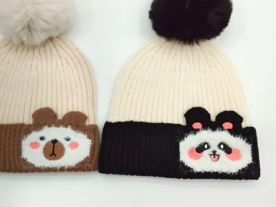 Zwei gerippte Strickmützen mit flauschigen Bommeln: beige Mütze mit Bären-Applikation und cremefarbenem Pompon sowie weiße Mütze mit Panda-Patch und schwarzem Bommel