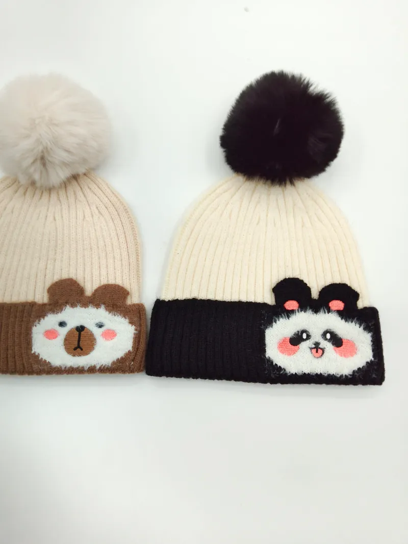 Zwei gerippte Strickmützen mit flauschigen Bommeln: beige Mütze mit Bären-Applikation und cremefarbenem Pompon sowie weiße Mütze mit Panda-Patch und schwarzem Bommel