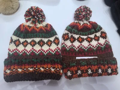 Zwei identische Fair-Isle-Strickmützen mit Bommel in Braun, Orange, Grün, Rot und Creme auf weißer Oberfläche nebeneinander
