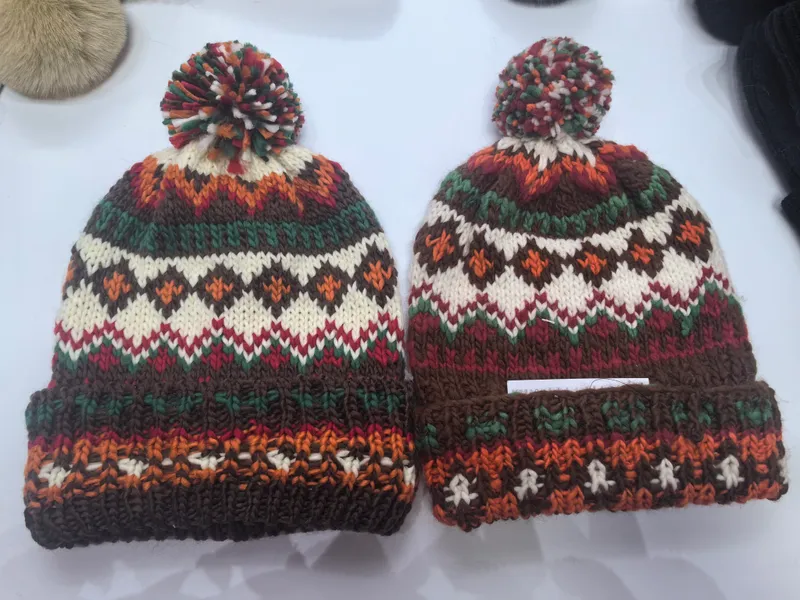 Zwei identische Fair-Isle-Strickmützen mit Bommel in Braun, Orange, Grün, Rot und Creme auf weißer Oberfläche nebeneinander