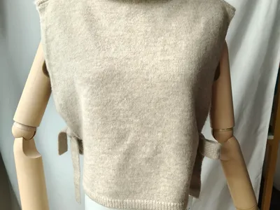 Beige ärmellose Rollkragen-Strickweste mit seitlichem Bindedetail auf einer Modelpuppe, weicher Wollmix
