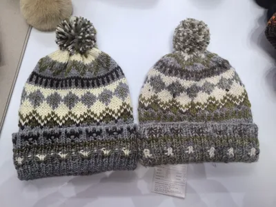 Zwei grob gestrickte Fair-Isle-Wintermützen mit Pompon in Grauton, Olivgrün und Cremeweiß mit Umschlagbündchen, paarweise präsentiert