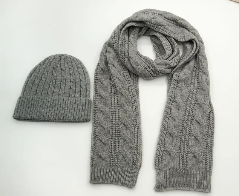 Passendes Winterset aus grauer Zopfstrickmütze und langem Schal im gleichen Flechtmuster, nebeneinander auf weißem Hintergrund