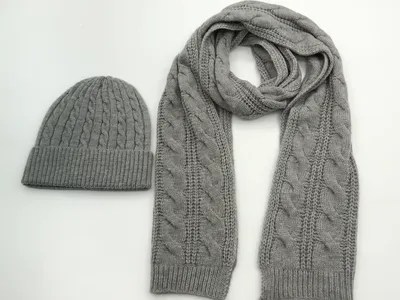 Passendes Winterset aus grauer Zopfstrickmütze und langem Schal im gleichen Flechtmuster, nebeneinander auf weißem Hintergrund