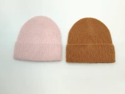 Zwei flauschige Wintermützen mit Umschlagbund nebeneinander, eine in zartem Rosa und eine in warmem Kamelbraun, Draufsicht auf weißem Untergrund
