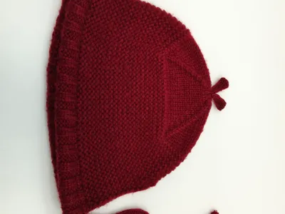 Handgestrickte Babymütze in kräftigem Dunkelrot mit Perlmuster-Textur, Rüschenkante, Kinnbändern zum Binden und charakteristischer Spitze, auf weißem Hintergrund liegend