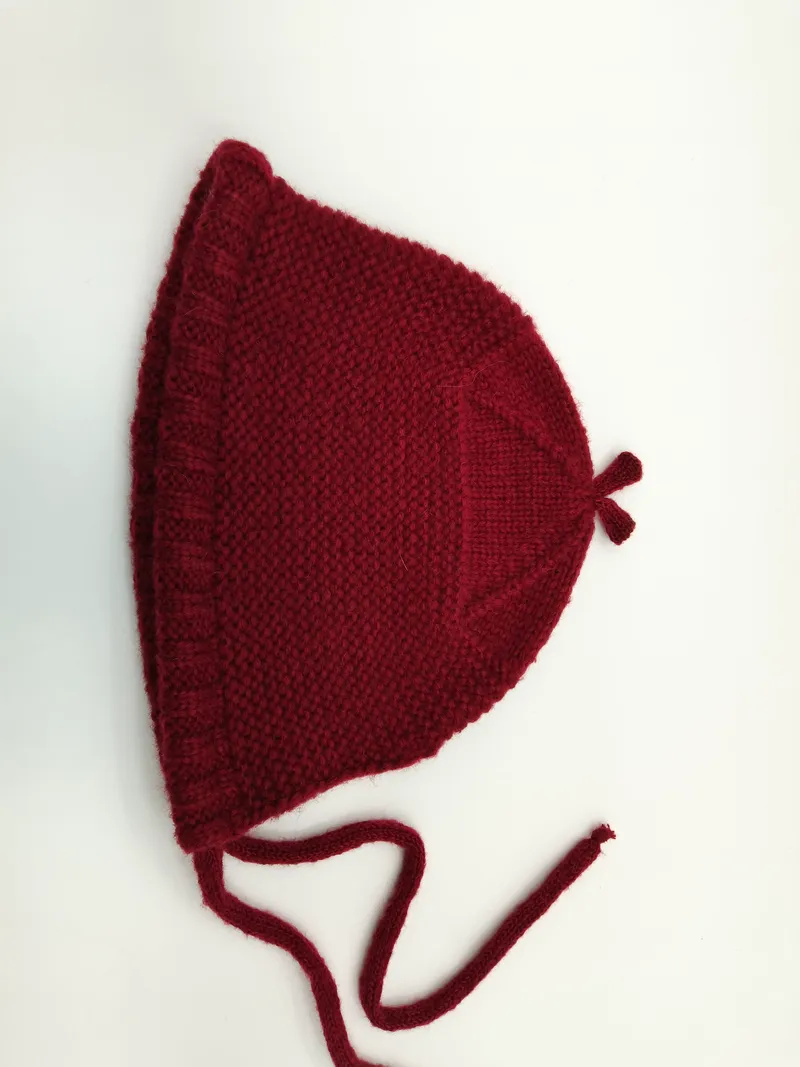 Handgestrickte Babymütze in kräftigem Dunkelrot mit Perlmuster-Textur, Rüschenkante, Kinnbändern zum Binden und charakteristischer Spitze, auf weißem Hintergrund liegend