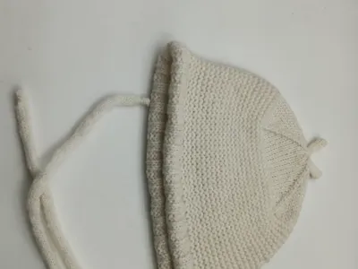 Cremeweiße handgestrickte Säuglingshäubchen mit Kinnbändern und feiner Perlstrick-Textur, auf hellem Untergrund liegend