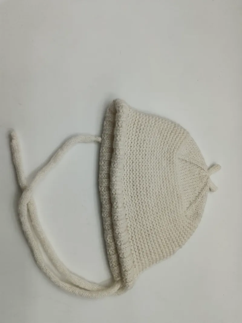 Cremeweiße handgestrickte Säuglingshäubchen mit Kinnbändern und feiner Perlstrick-Textur, auf hellem Untergrund liegend