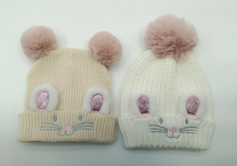 Zwei gestrickte Baby-Mützen in Creme und Weiß mit glitzernden Hasenohren, aufgestickten Schnurrhaaren und weichen pinken Kunstfell-Pompons oben