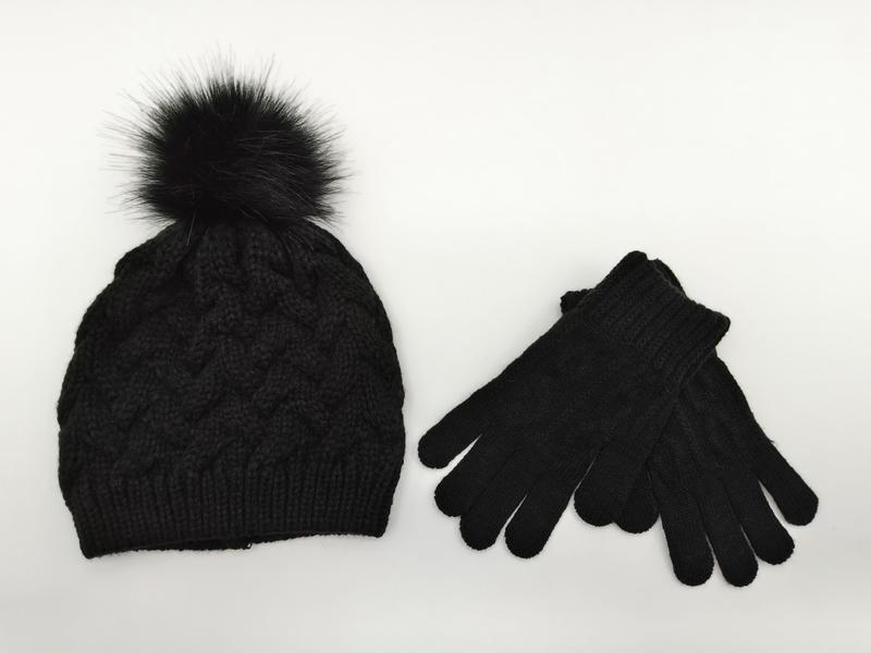 Schwarze Zopfstrick-Wintermütze mit fluffigem Kunstfell-Pompon und dazu passenden schwarzen Strickhandschuhen auf weißem Untergrund