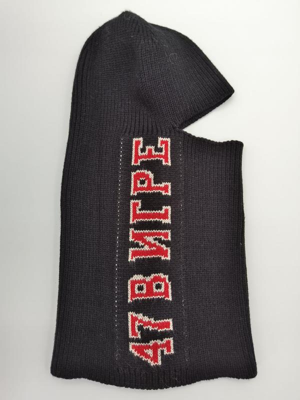 Schwarze Rippenstruktur-Skimaske flach präsentiert mit rot-weißem Varsity-Letter-Aufdruck auf dunklem Strickgewebe