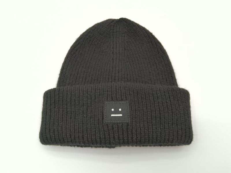 Schwarze Wintermütze in grobem Rippenstrick mit umgeschlagenem Bund und kleinem quadratischem besticktem Logo-Aufnäher auf weißem Hintergrund