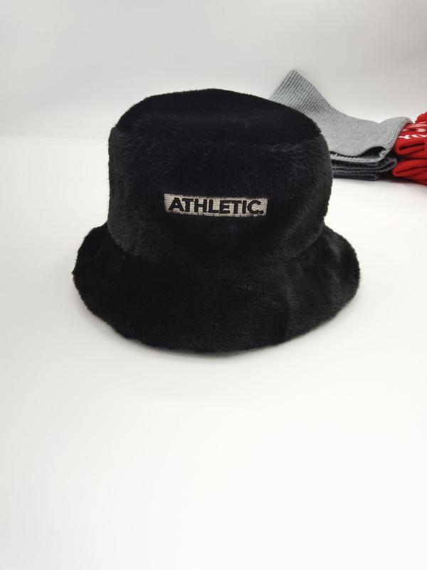 Schwarze flauschige Kunstfell-Fischermütze mit gesticktem Athletic-Etikett-Aufnäher, präsentiert auf weißer Oberfläche mit grauen und roten Accessoires