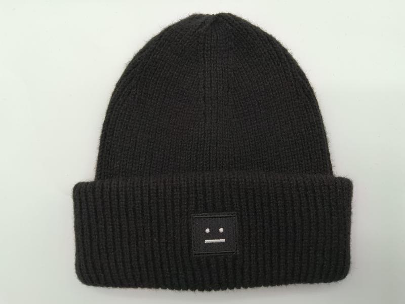 Schwarze fein gerippte Strickmütze mit umgeschlagenem Rand und kleinem besticktem Motiv-Patch auf weißem Hintergrund