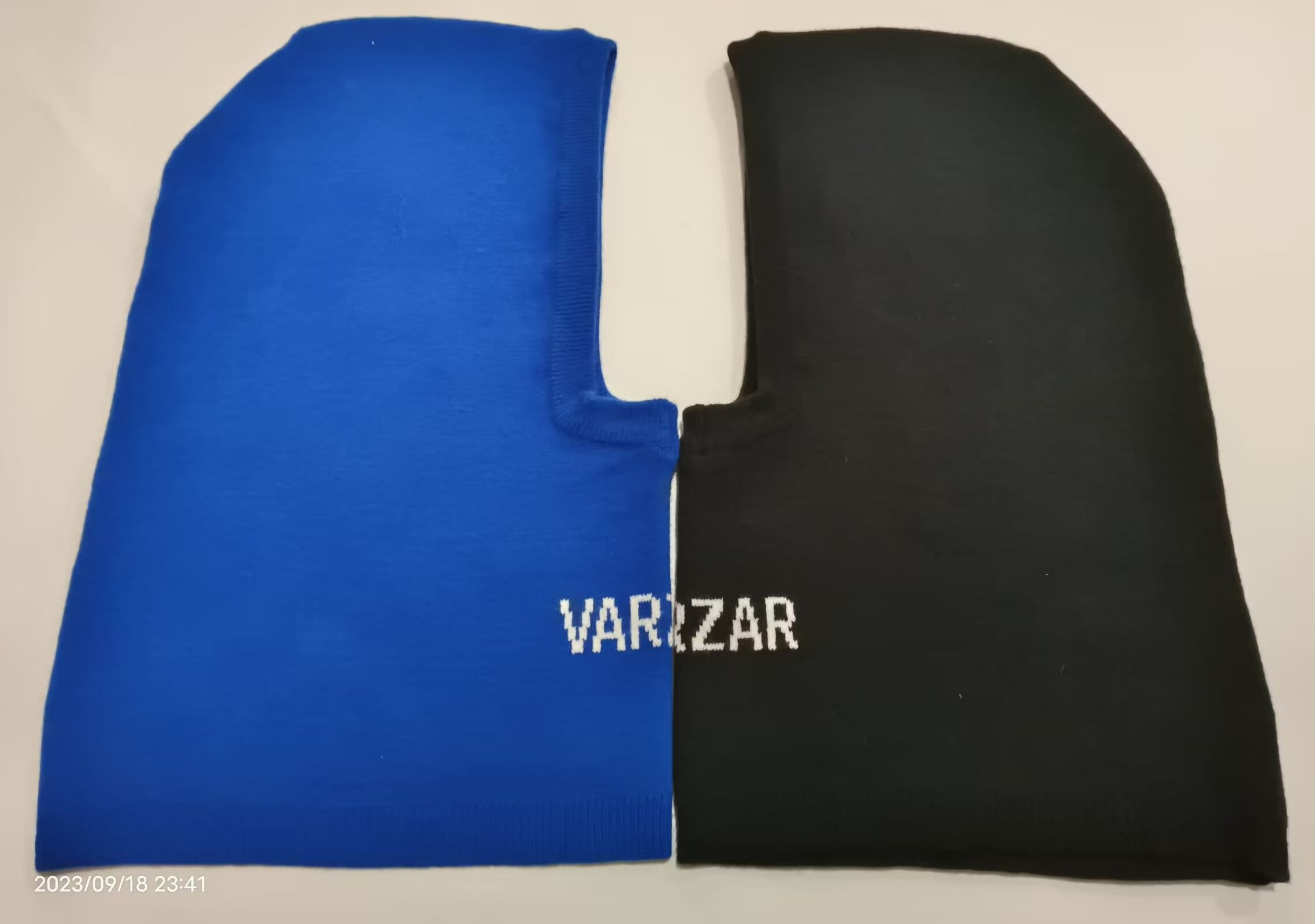 Blau-schwarze zweifarbige gestrickte Sturmhaube flach liegend mit Gesichtsausschnitt und Logoprint auf der schwarzen Seite