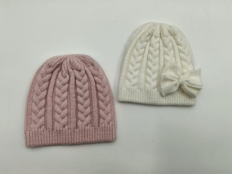 Zwei Wintermützen mit klassischem Zopfstrickmuster – eine in gedämpftem Rosa, eine in Cremeweiß mit seitlicher Schleifenverzierung